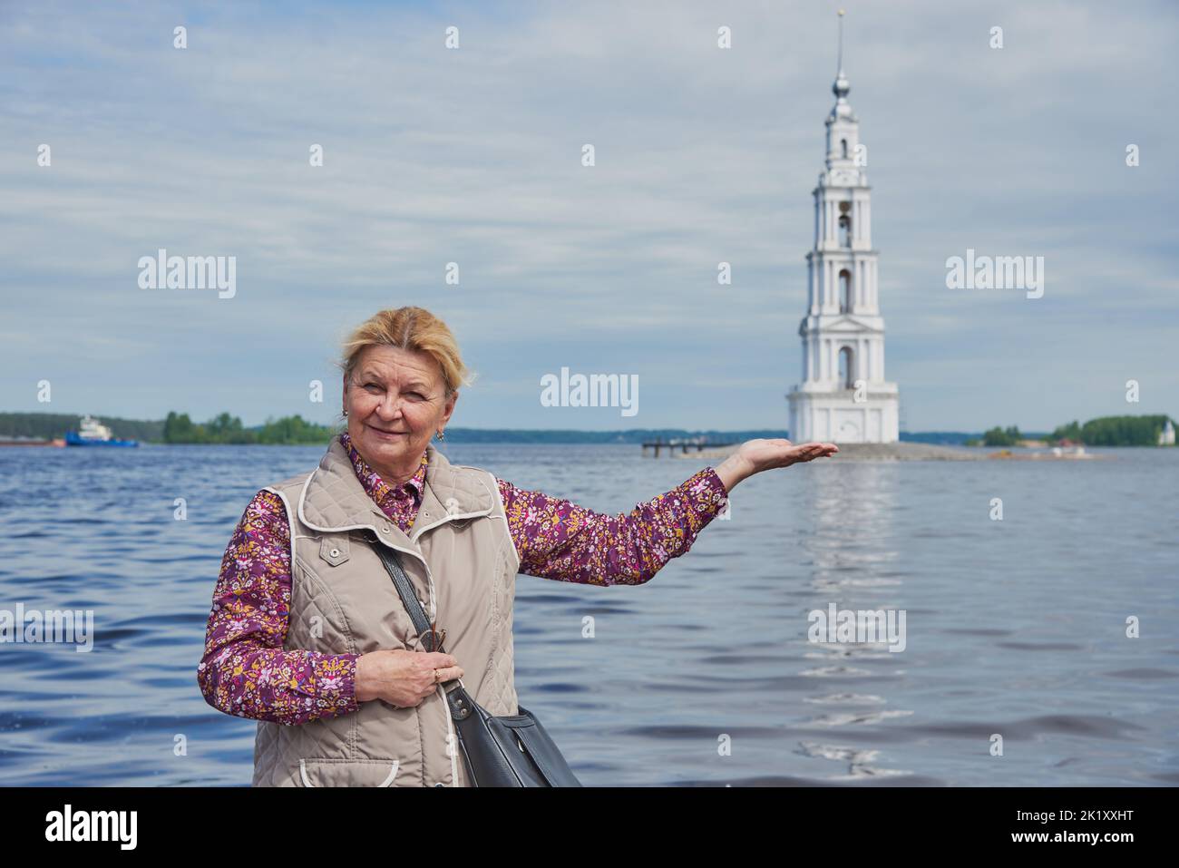 An elderly tourist poses against the background of the bell tower and pretends to hold the tower in the palm of her hand.  Stock Photo