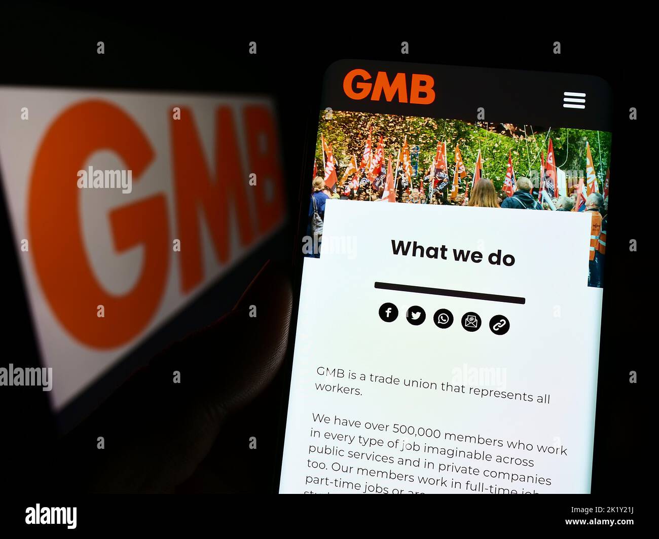 Person holding cellphone with webpage of British trade union GMB on screen in front of logo. Focus on center of phone display. Stock Photo