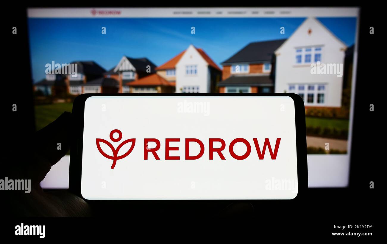 Person holding smartphone with logo of British housebuilding company Redrow plc on screen in front of website. Focus on phone display. Stock Photo