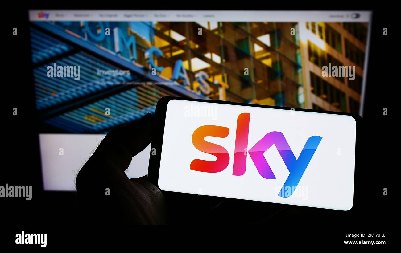 Person holding mobile phone with logo of British media company Sky Group Limited on screen in front of business web page. Focus on phone display. Stock Photo