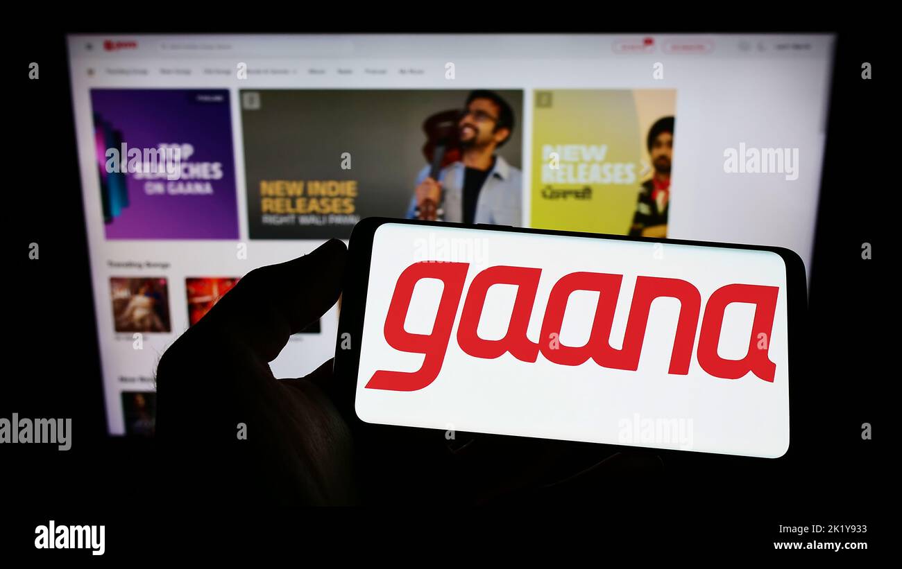 Person holding mobile phone with logo of music streaming company Gamma Gaana Ltd. on screen in front of business web page. Focus on phone display. Stock Photo