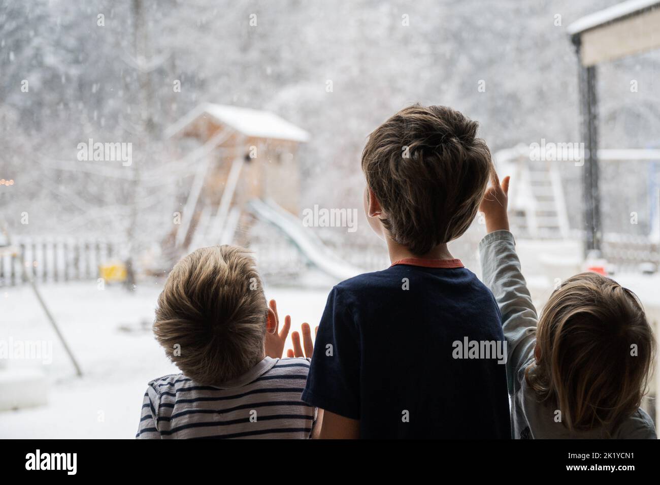 View from behind of three children, siblings, looking out the window into a beautiful winter nature with snow falling down the sky. Stock Photo