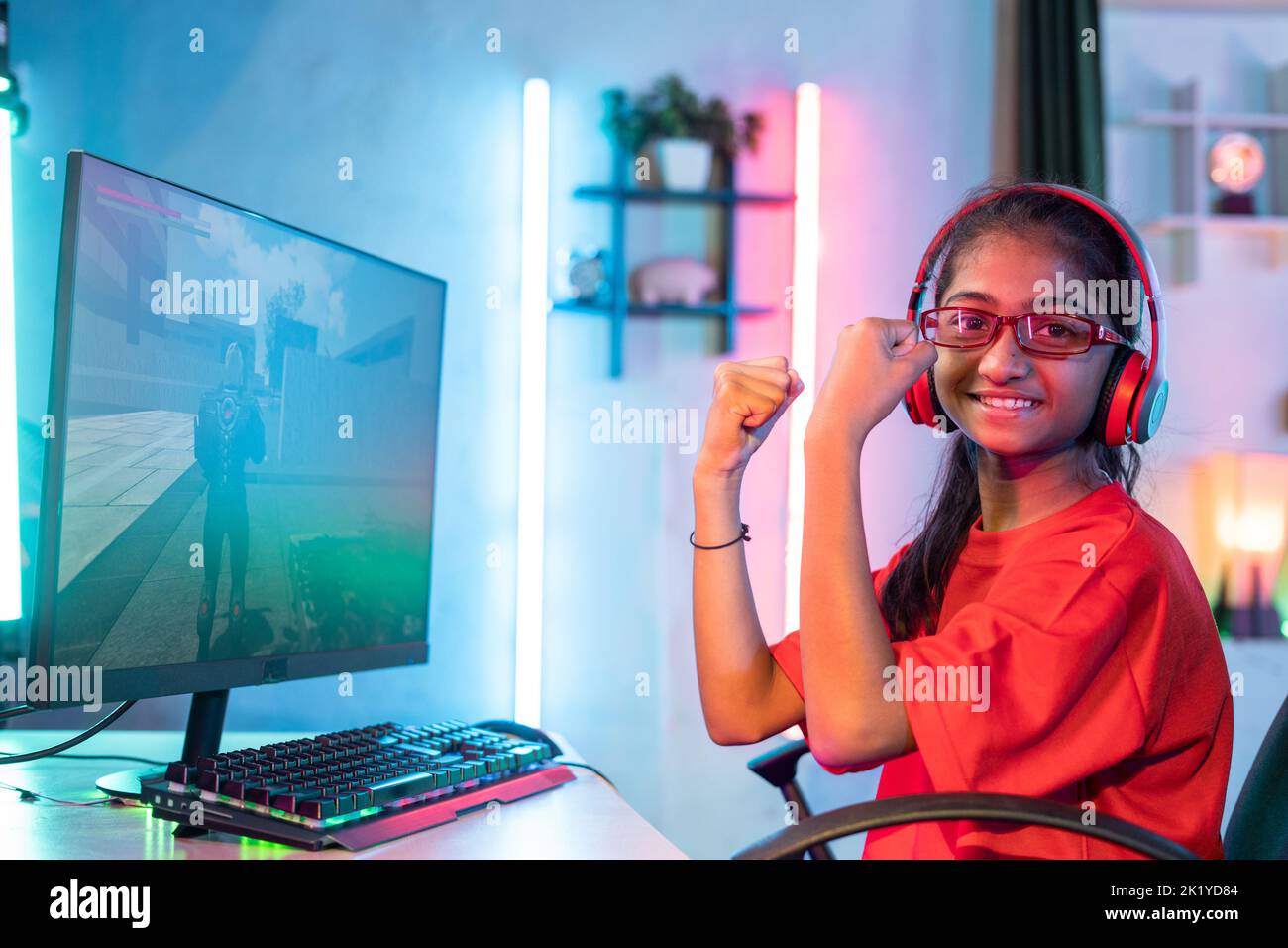 Excited girl with headset celebrating by raising hands while playing live video game on computer by looking at camera - concept of social media Stock Photo