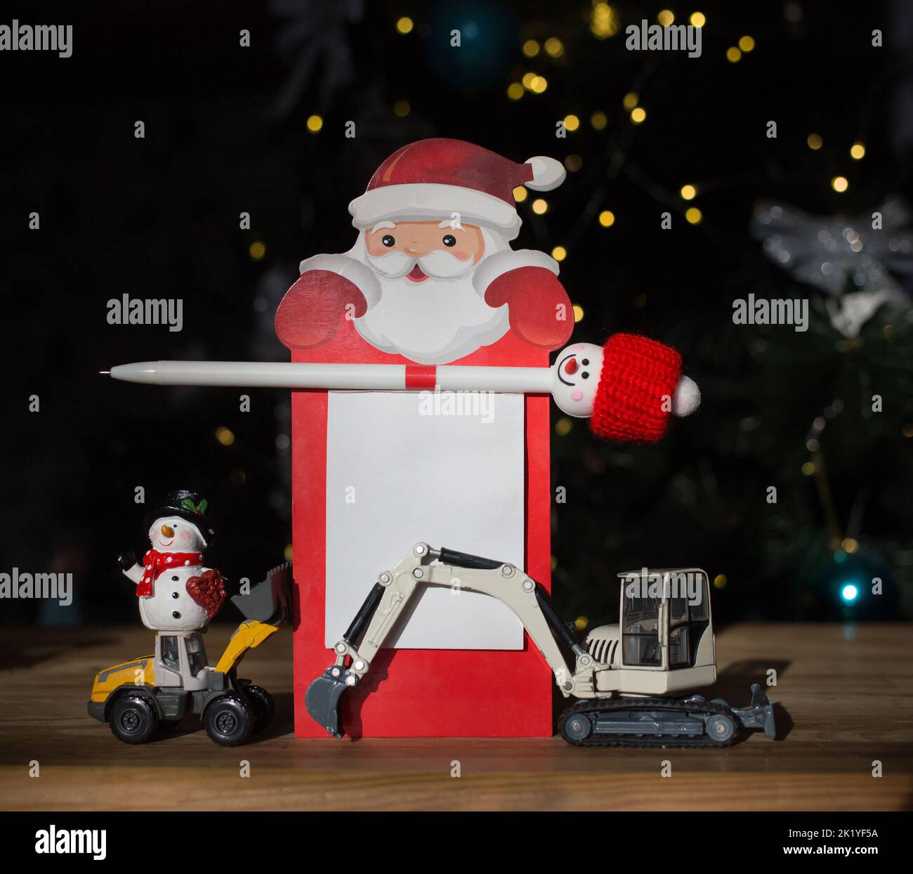 2 models of toy excavators, snowman, notebook with Santa Claus, pen on background of Christmas tree. concept of New Year's business greetings for cons Stock Photo