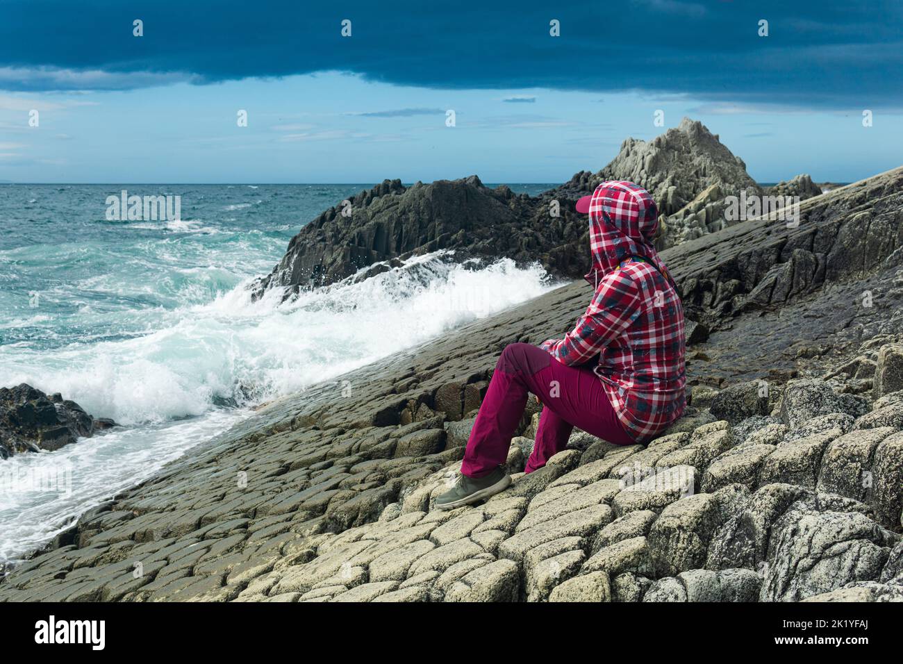 girl in a jacket with a hood sits on the shore of a stormy sea, on a ...