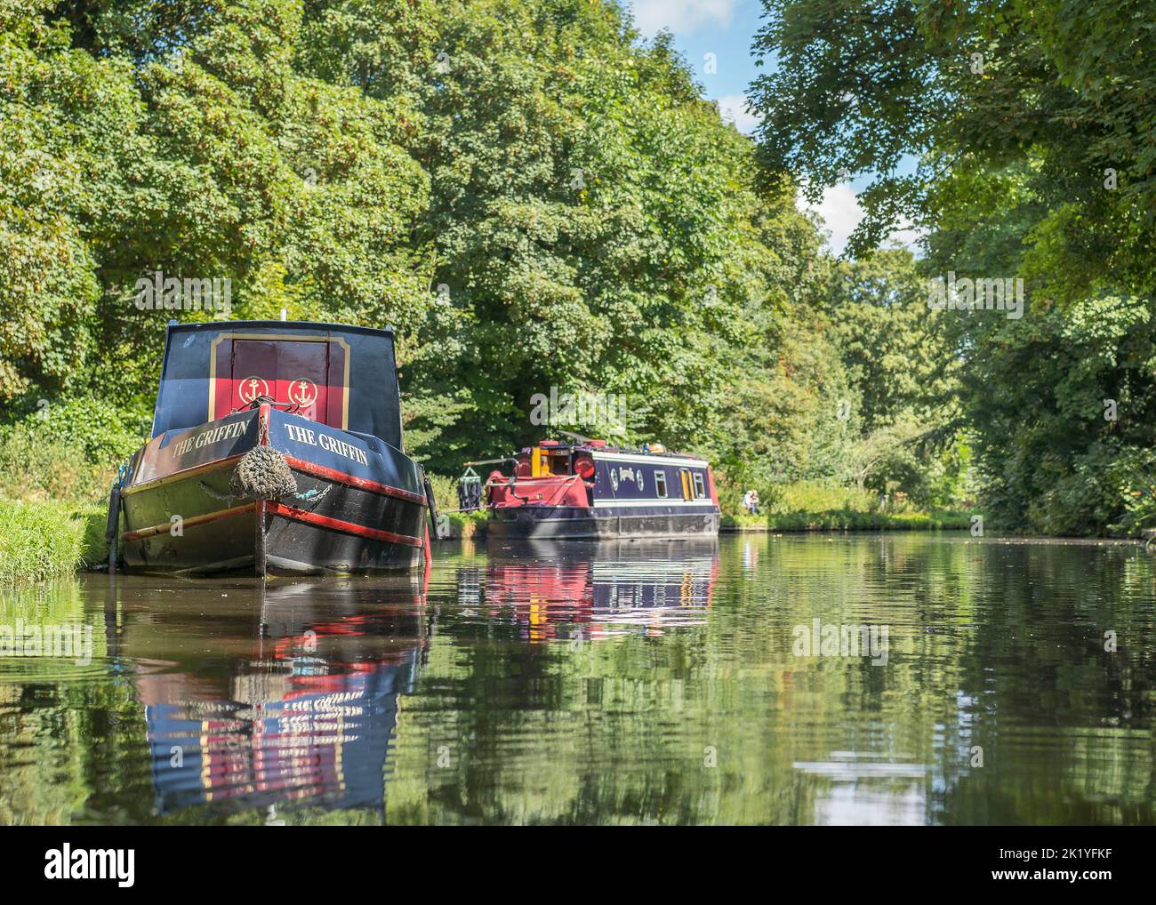Low angle view of two canal boats moored at the side of a wide canal with their reflections in the water on a sunny day. Stock Photo