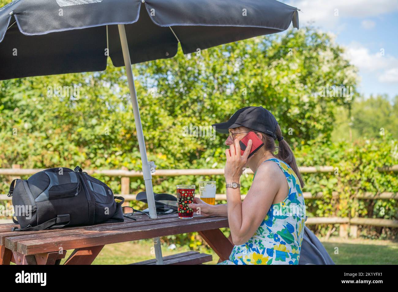 Side view of woman sat drinking in a pub garden on a sunny day talking on her mobile phone, sheltering under a garden brolly for sun protection. Stock Photo
