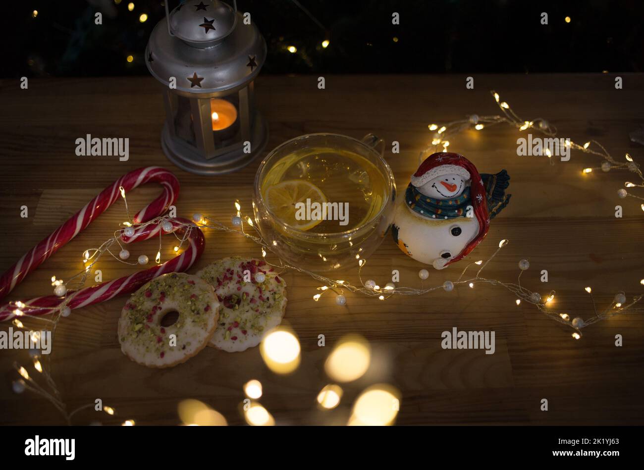 Cozy atmospheric photo of Christmas theme. transparent cups with tea and lemon slices, souvenir snowman, cookies, glowing garland lights, lollipops, b Stock Photo