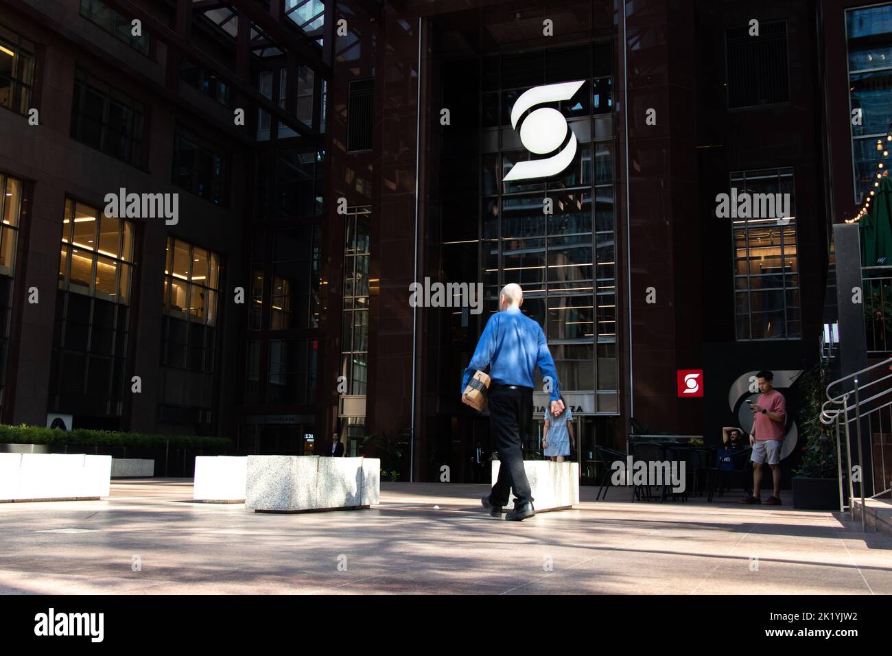 A businessman walks towards Scotia Plaza in downtown Toronto; the Scotiabank logo is seen illuminated in the shade, as the sun glares down. Stock Photo