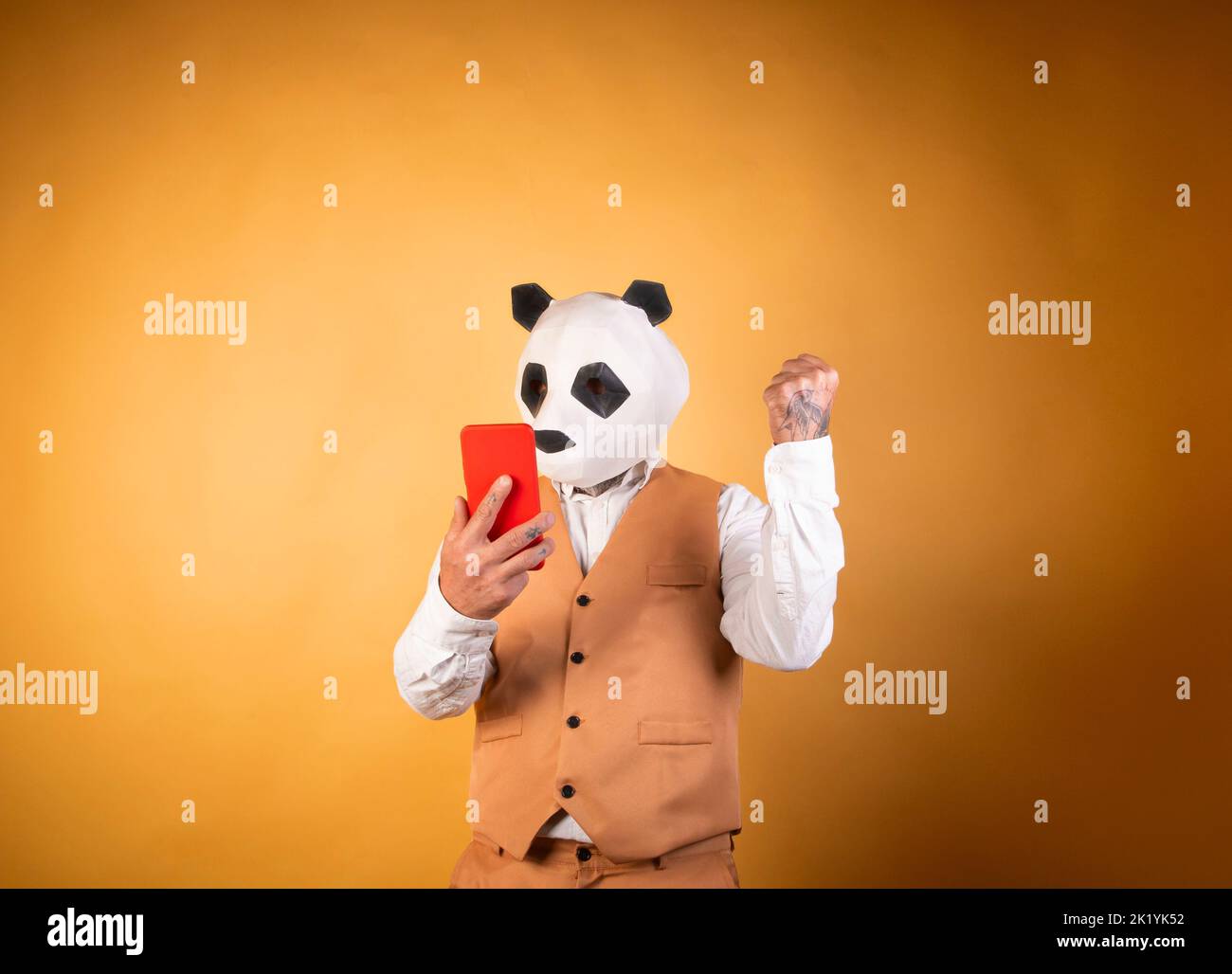 Man in panda bear mask on yellow background using smartphone. Stock Photo