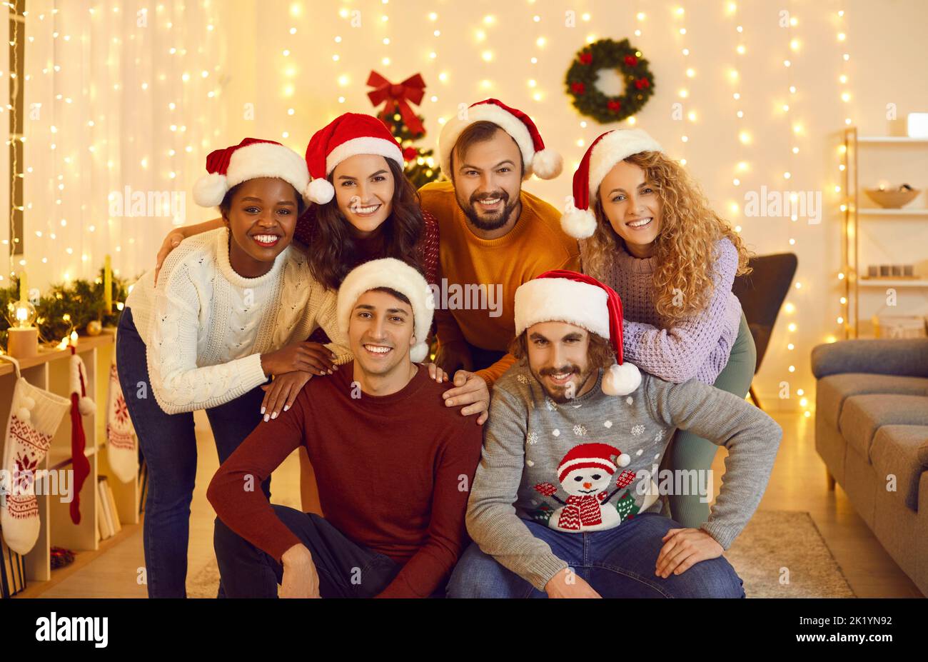 Portrait of cheerful young friends in Santa hats posing in cozy living room with Christmas decor. Stock Photo