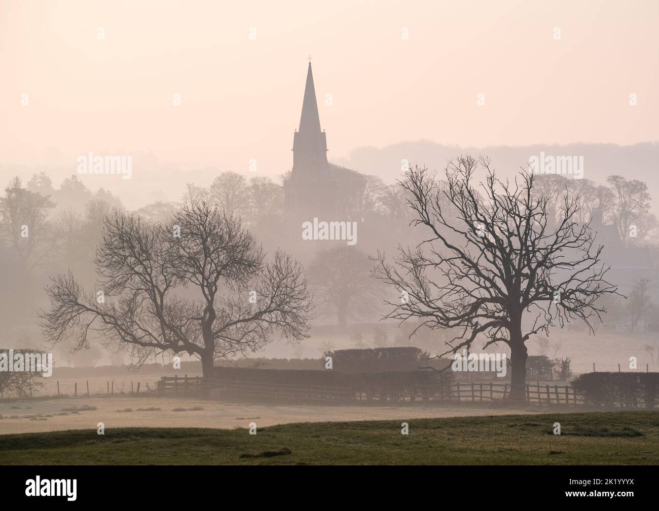St Barnabas Church in Weeton is visible through the mist on a calm spring morning, framed by the skeletal outlines of two leafless trees. Stock Photo