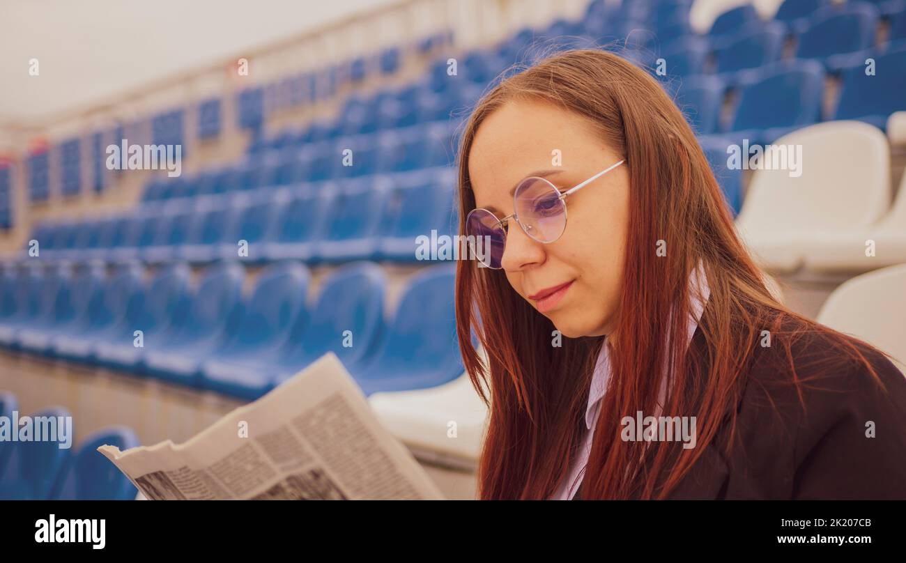 A woman in glasses and a business suit reads a newspaper while sitting at the stadium. Stock Photo