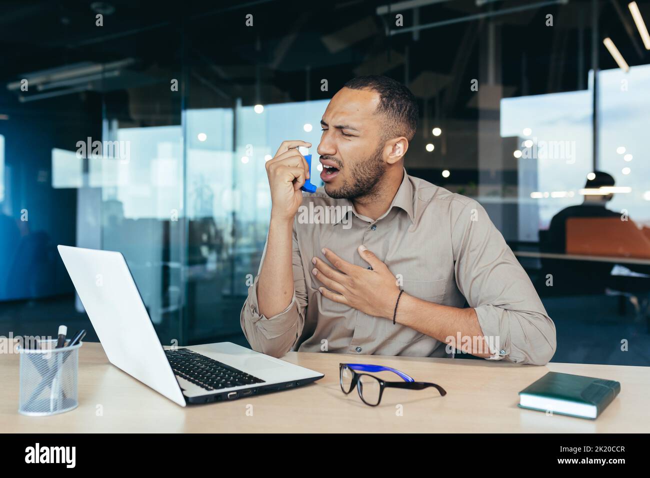 Sick office worker with asthma having trouble breathing, man using inhaler to relieve breathing, hispanic man working inside modern office building, mixed race man with laptop Stock Photo