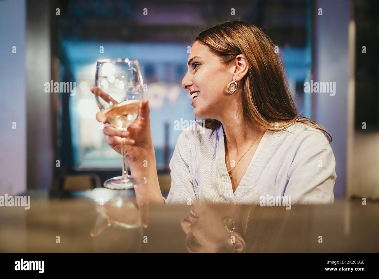 Portrait of a long haired young adult woman holding a glass of white wine sitting at restaurant counter - view from the counter - people and alcohol n Stock Photo