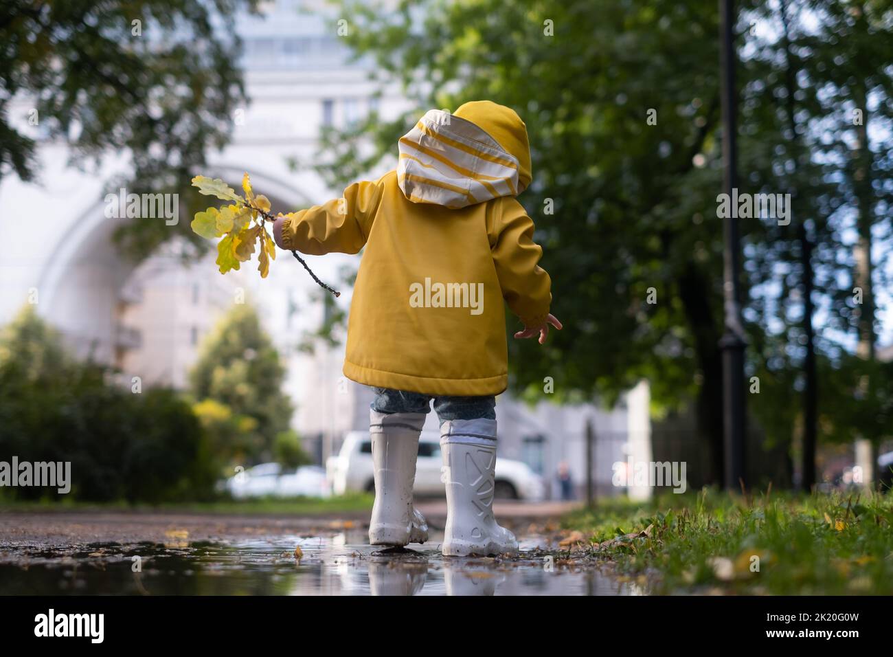 Cute little caucasian girl in yellow jacket, blue jeans and white rubber boots is running over a puddle Stock Photo
