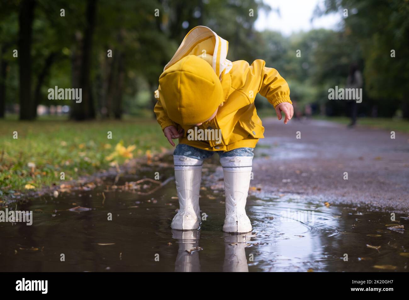 Cute little caucasian girl in yellow jacket, blue jeans and white rubber boots is running over a puddle Stock Photo