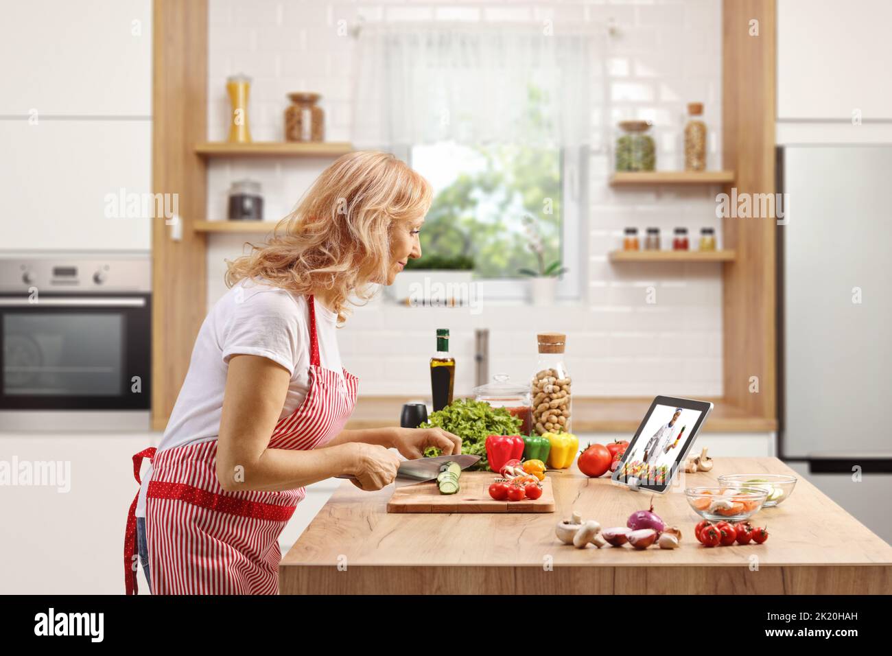 Mature woman cutting vegetables and watching a video on a tablet in a kitchen Stock Photo