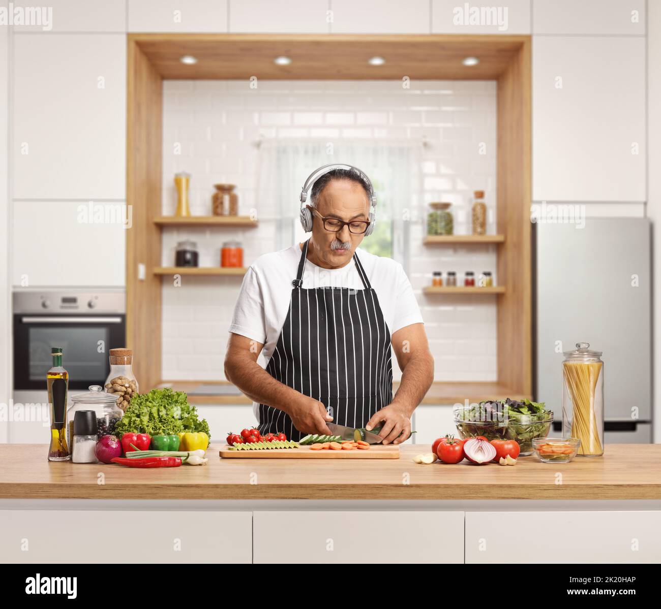 Mature man cutting vegetables on a wooden counter and listening to music with headphones inside a kitchen Stock Photo