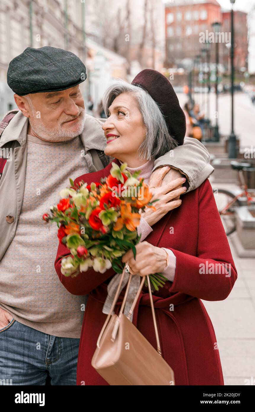 Elderly senior love couple. Old retired man woman together on romantic date.Aged husband wife walking on city street with flowers.Stylish elder huggin Stock Photo