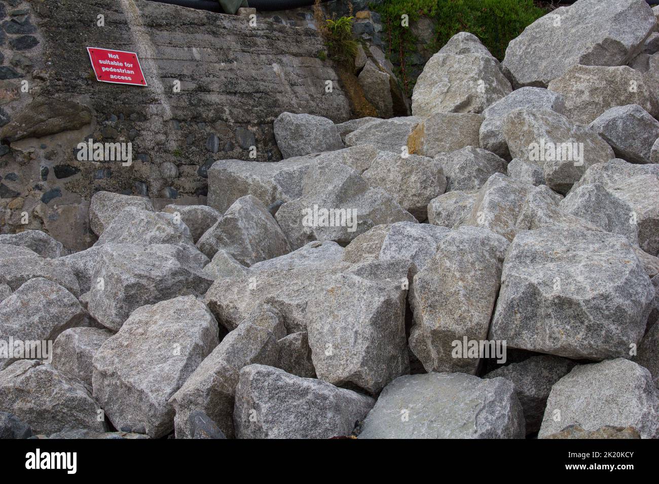 Rock armour transported to Coverack Bay, Cornwall.  To reinforce the sea wall against storm damage and erosion. Ironic sign warning pedestrians. Stock Photo
