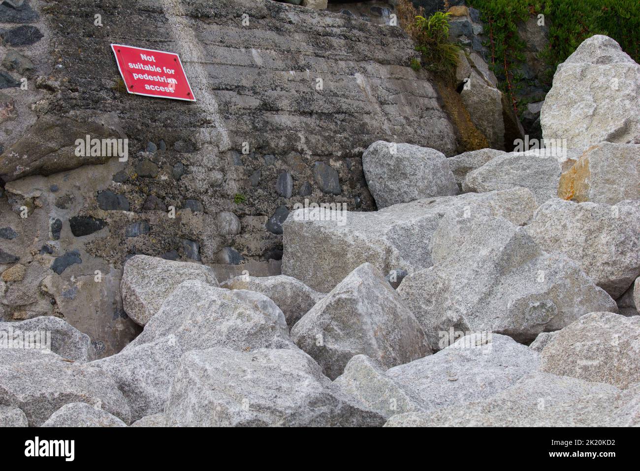 Rock armour transported to Coverack Bay, Cornwall.  To reinforce the sea wall against storm damage and erosion. Ironic sign warning pedestrians. Stock Photo