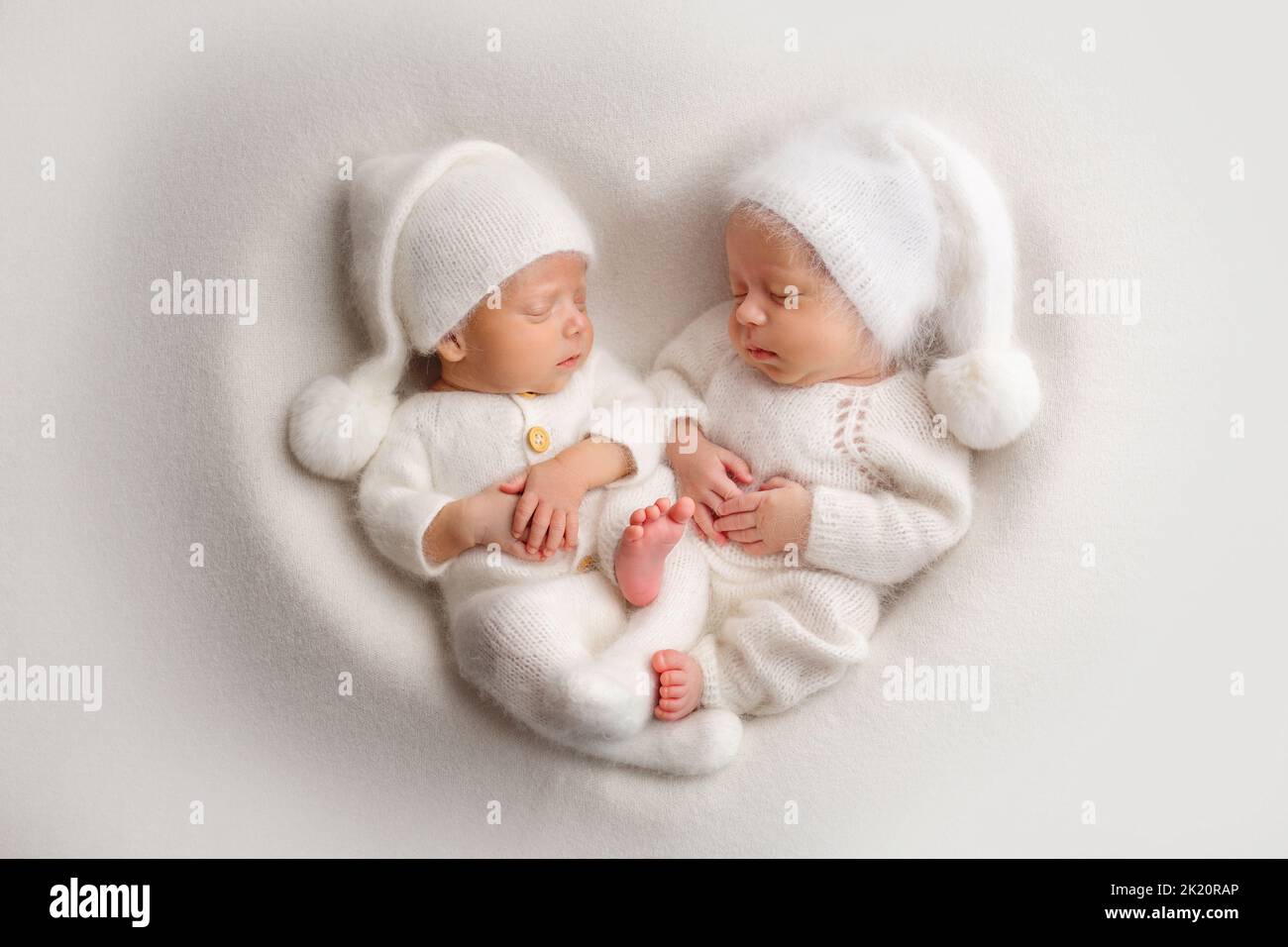 Tiny newborn twin boys in white bodysuits in white caps. Newborn twins