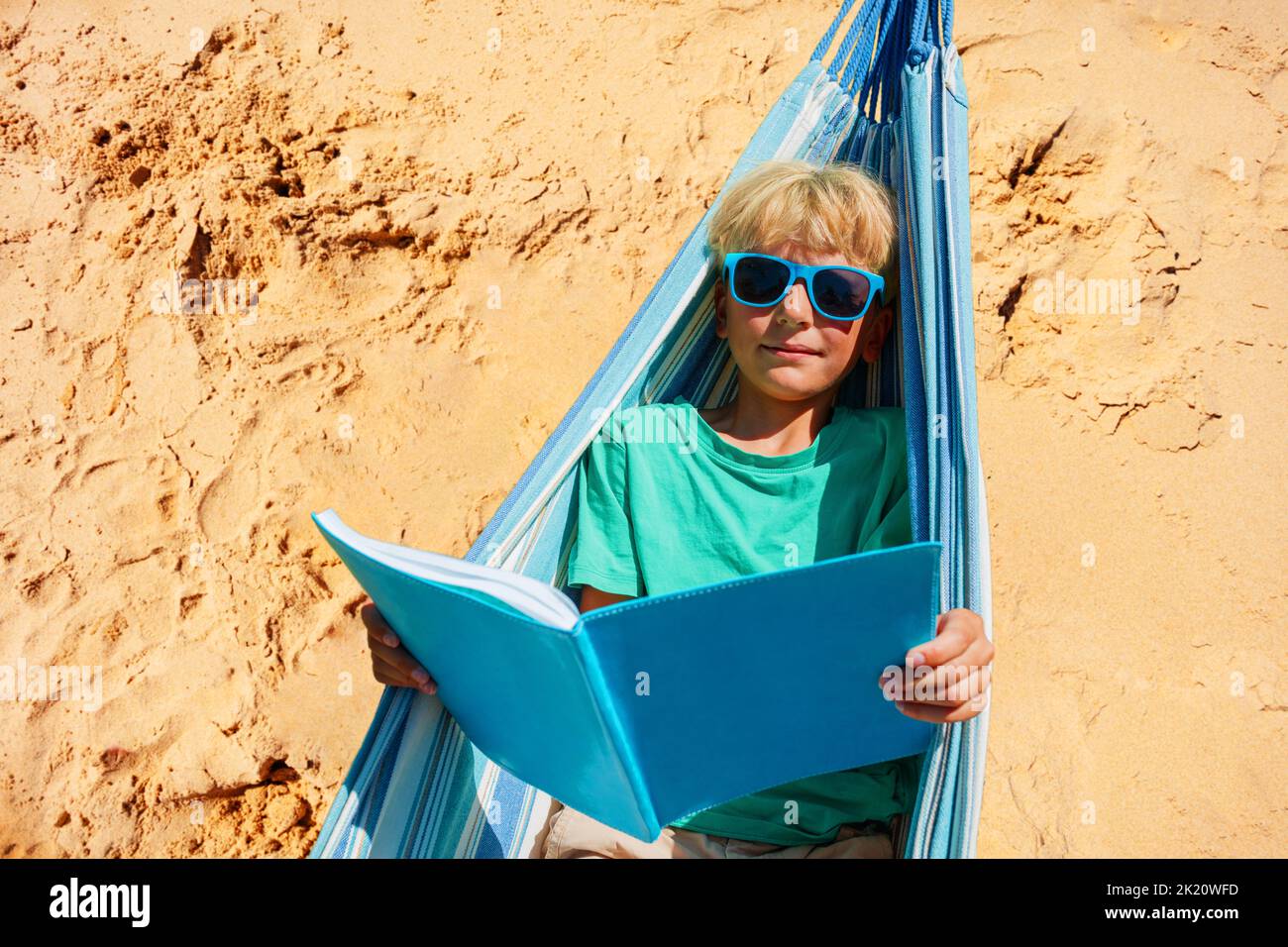 Boy with sunglasses lay in hammock and read a book at beach Stock Photo