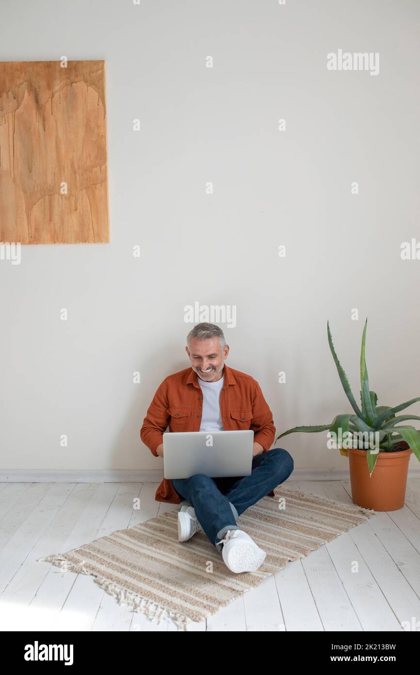 Man in terracotta shirt sitting on the floor with a laptop in hands Stock Photo