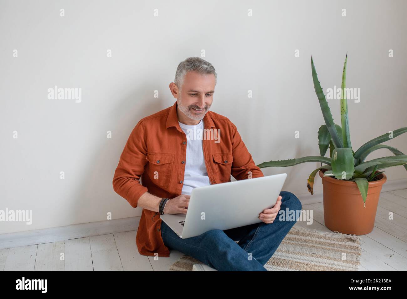 Man in terracotta shirt sitting on the floor with a laptop in hands Stock Photo
