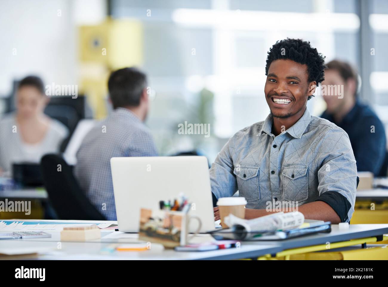 Its been a fun ride getting to where I am. Portrait of a designer sitting at his desk working on a laptop with colleagues in the background. Stock Photo