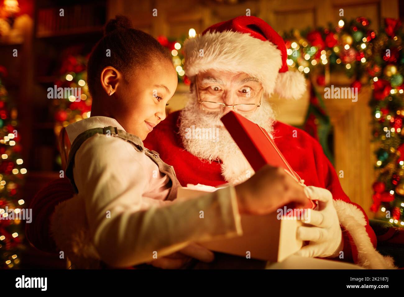 Side view portrait of cute black girl opening Christmas present with traditional Santa Claus, magic light Stock Photo