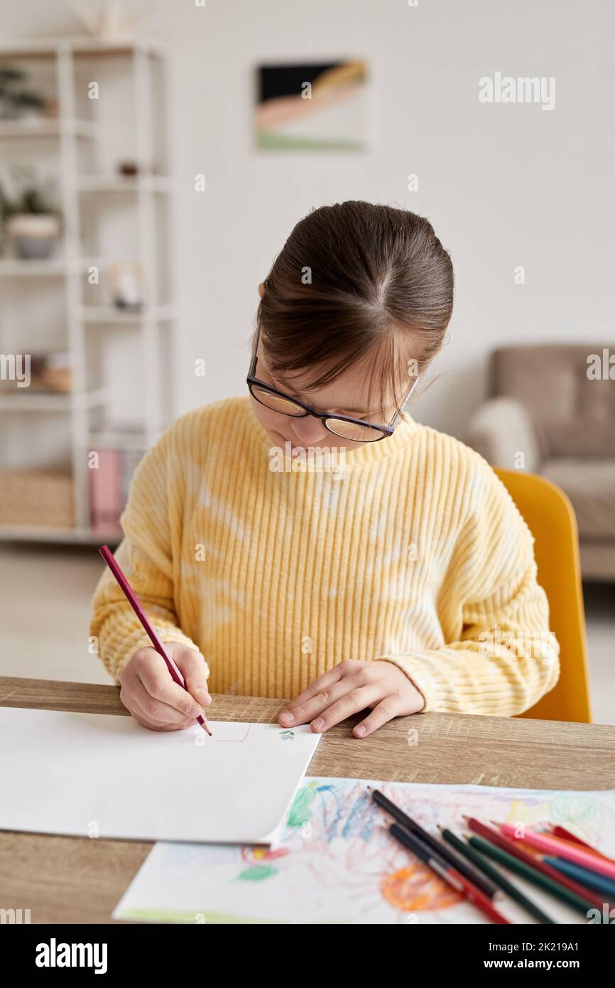 Vertical portrait of teenage girl with Down syndrome drawing pictures at table in cozy room Stock Photo