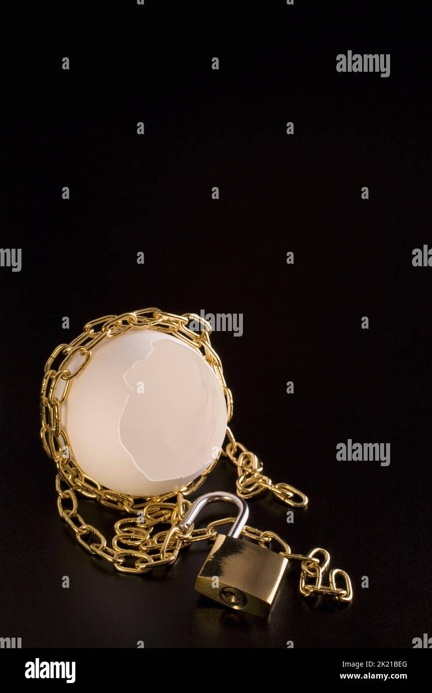 Close-up of opened brass padlock and cracked opened egg shell wrapped with a gold chain on black glossy background, Studio Composition, Quebec, Canada Stock Photo