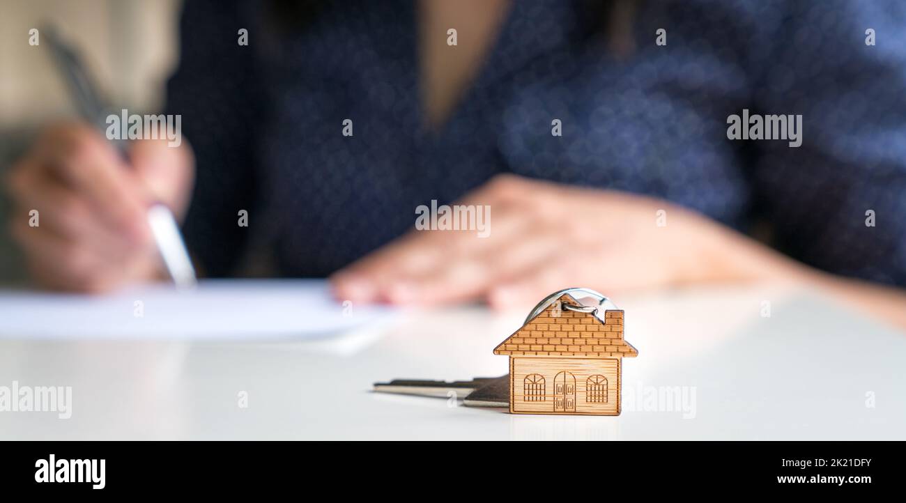 House shaped keychain with woman signing sale and purchase agreement at the background. Stock Photo
