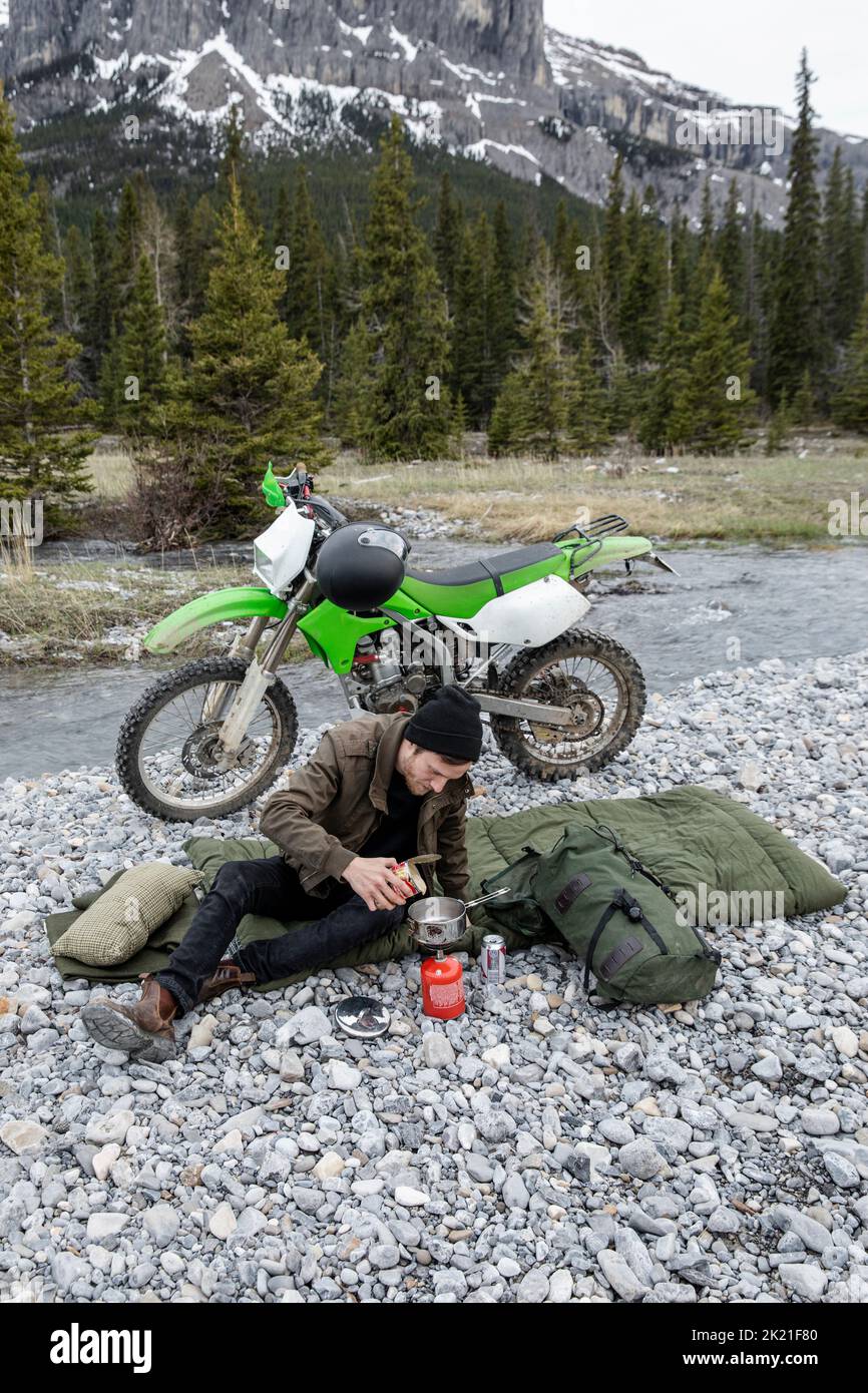Man heating canned food with camping stove at dirt bike below mountain