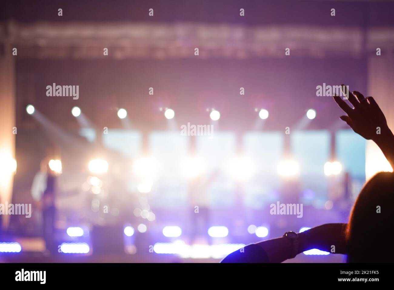 Defocus silhouette of one woman raise hand up in music concert with purple and white color spotlight on stage background. Out of focus. Stock Photo
