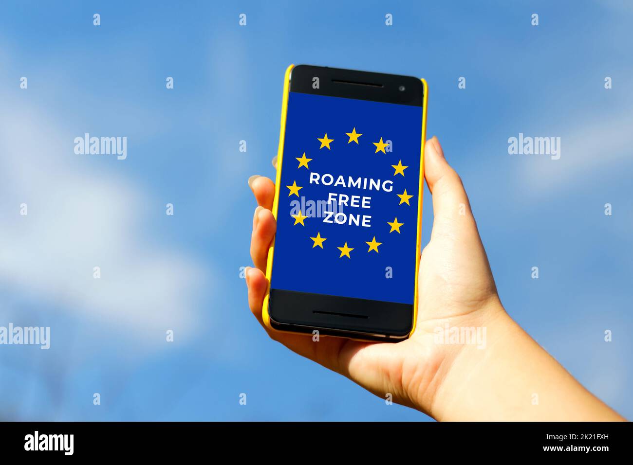 Roaming Free Euro Zone. Woman hand holding black smartphone in yellow plastic case on blue sky nature background, sunny day. Black screen. Female hand Stock Photo