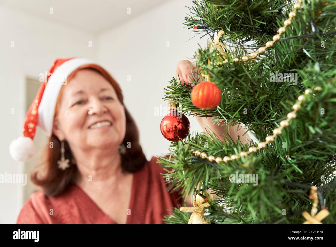 Happy senior hispanic woman smiling while decorating a Christmas tree at home wearing a red Santa Claus beanie. Selective focus in the foreground, a h Stock Photo