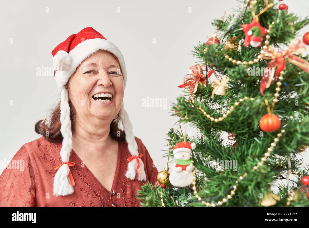 Happy senior hispanic woman laughing while decorating her Christmas tree at home, wearing a red Santa Claus beanie with white braids. The joy of the h Stock Photo
