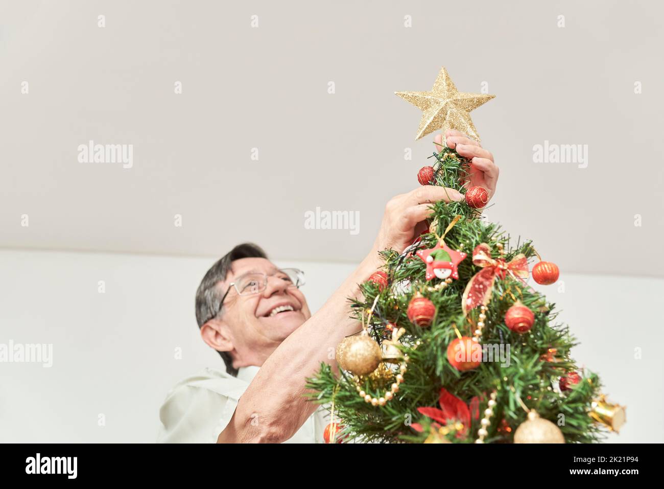 Happy senior latin man smiling as he puts a gold star on the top of his Christmas tree, the joy of spending the holidays at home. Stock Photo