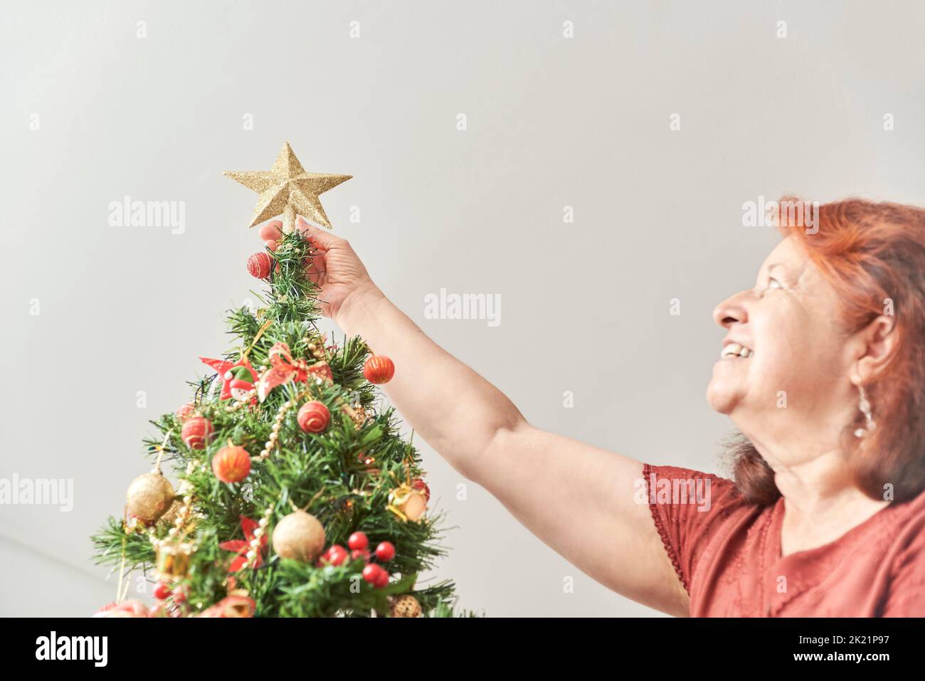 Happy mature latin woman smiling as she puts a gold star on the top of her Christmas tree, the joy of spending the holidays at home. Stock Photo