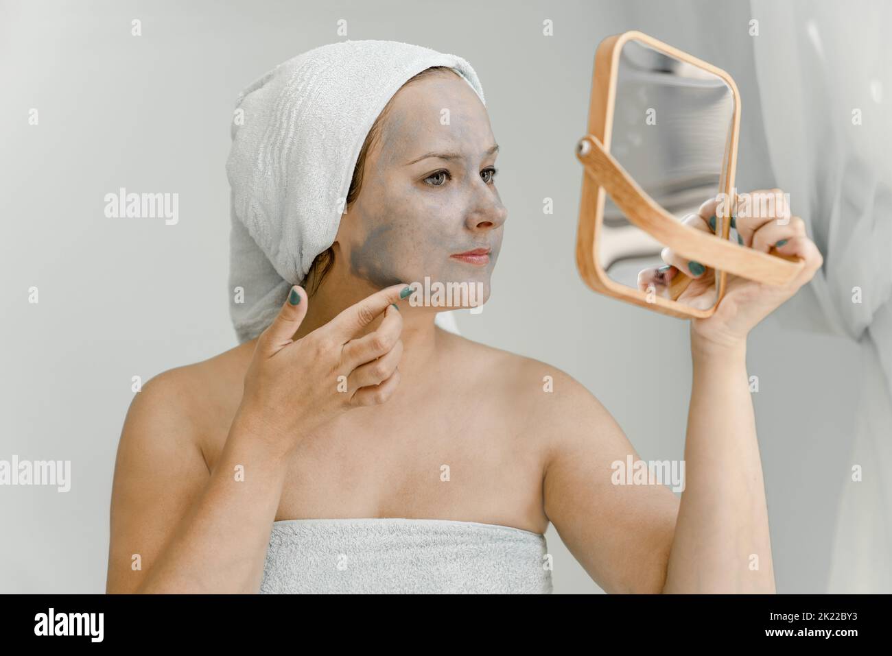 Young woman puts gray cosmetic clay mask on face while looking in mirror, her hair and body wrapped in towel. Close-up Stock Photo