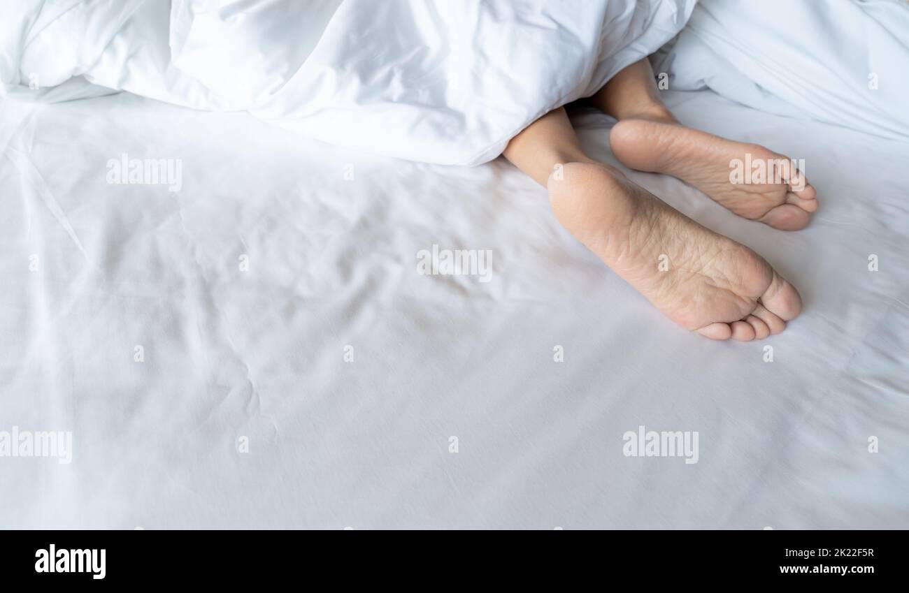 Woman barefoot on bed under white linen blanket in hotel or home bedroom. Healthy sleep and relaxation concept. Lazy Sunday morning. Bare feet Stock Photo