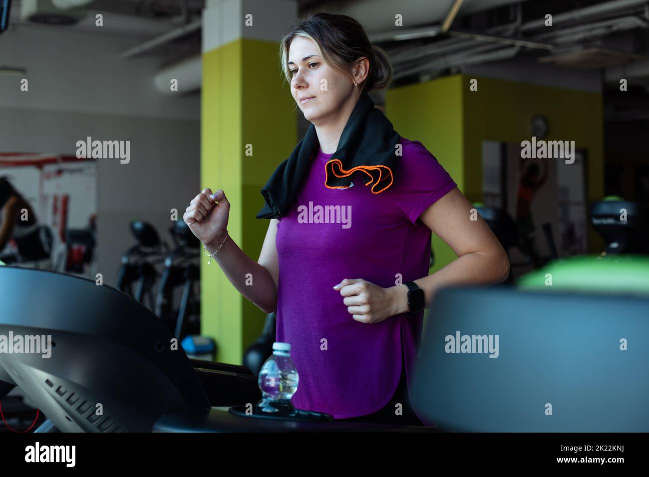 Overweight woman with pleasure run on treadmill at fitness gym with towel hanging around neck, side view. Young plus size girl do cardio workout on Stock Photo