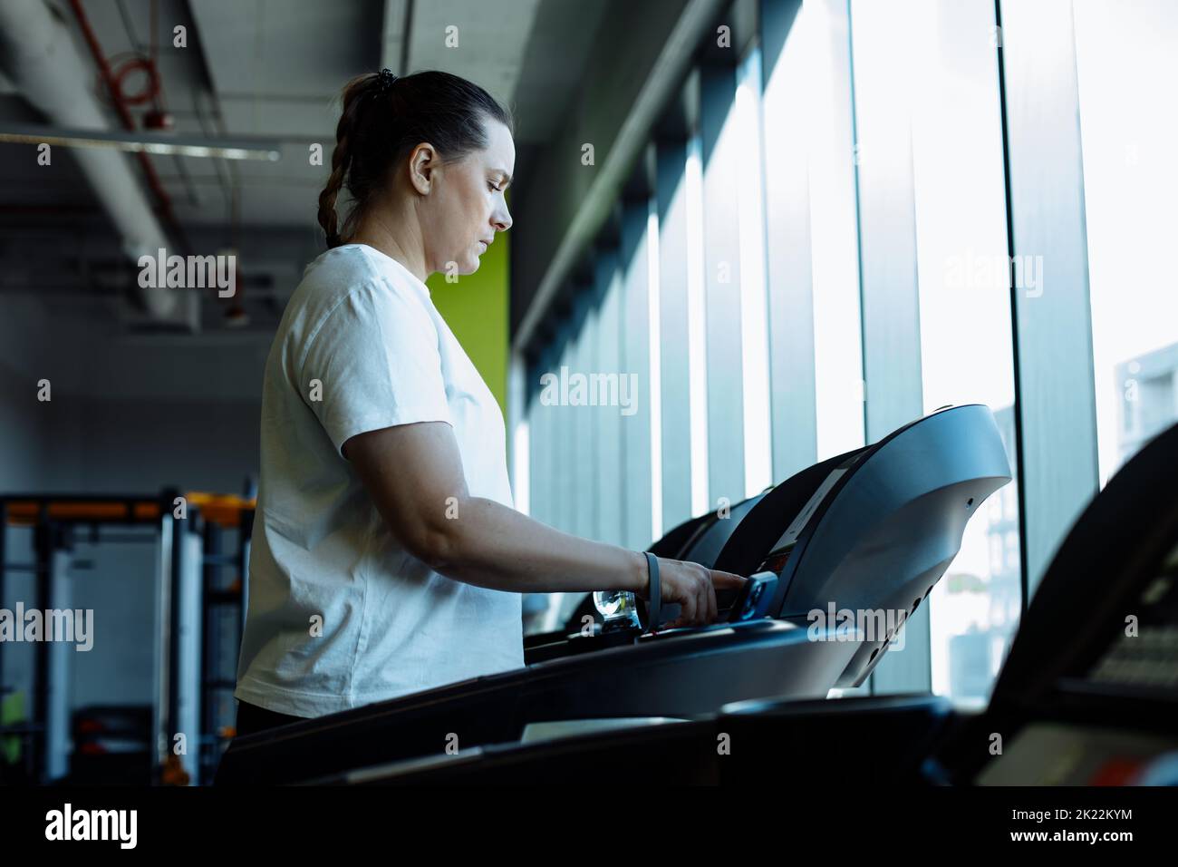 Adult plus size woman run on treadmill at fitness gym, side view. Middle aged lady set up speed mode on sport equipment for cardio workout. Weight Stock Photo