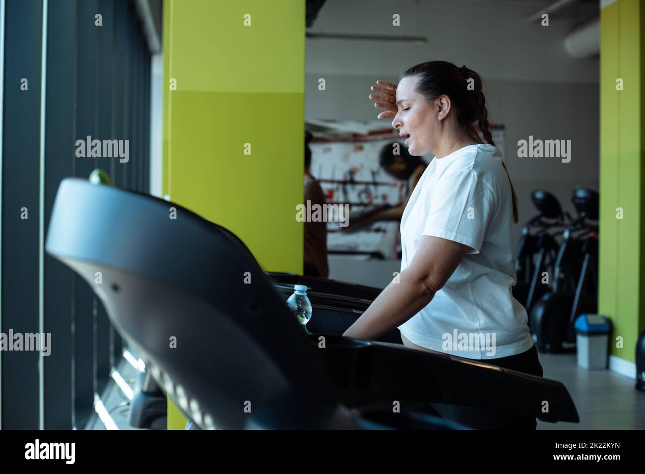 Tired plus size woman run on treadmill at fitness gym wiping sweat from face by hand, side view. Middle aged lady do cardio workout on exercise Stock Photo