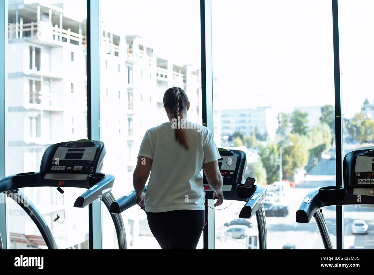 Fat woman alone run on treadmill at fitness gym and look out window at transport traffic and city, back view. Overweight lady do cardio workout on Stock Photo