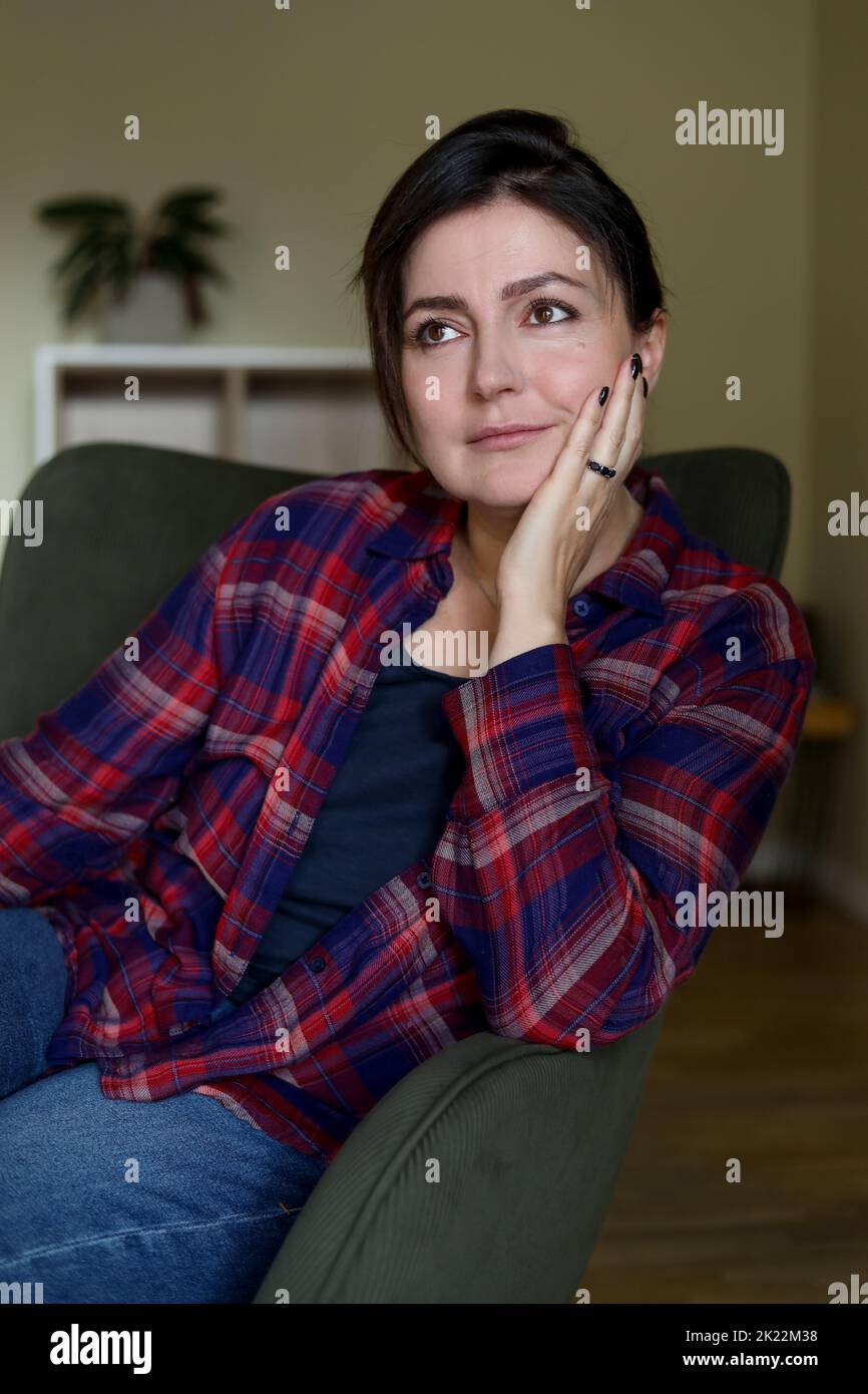 Beautiful brunette woman relax in comfortable armchair in home room and thoughtfully look away. Adult female people enjoying quiet lifestyle indoor Stock Photo