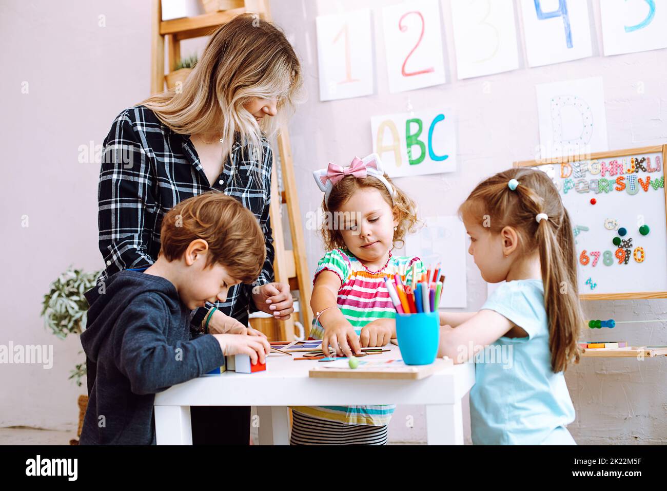 Group of kids playing together with educational toys in playroom of kindergarten. Young educator sitting with children at table on floor and help to Stock Photo